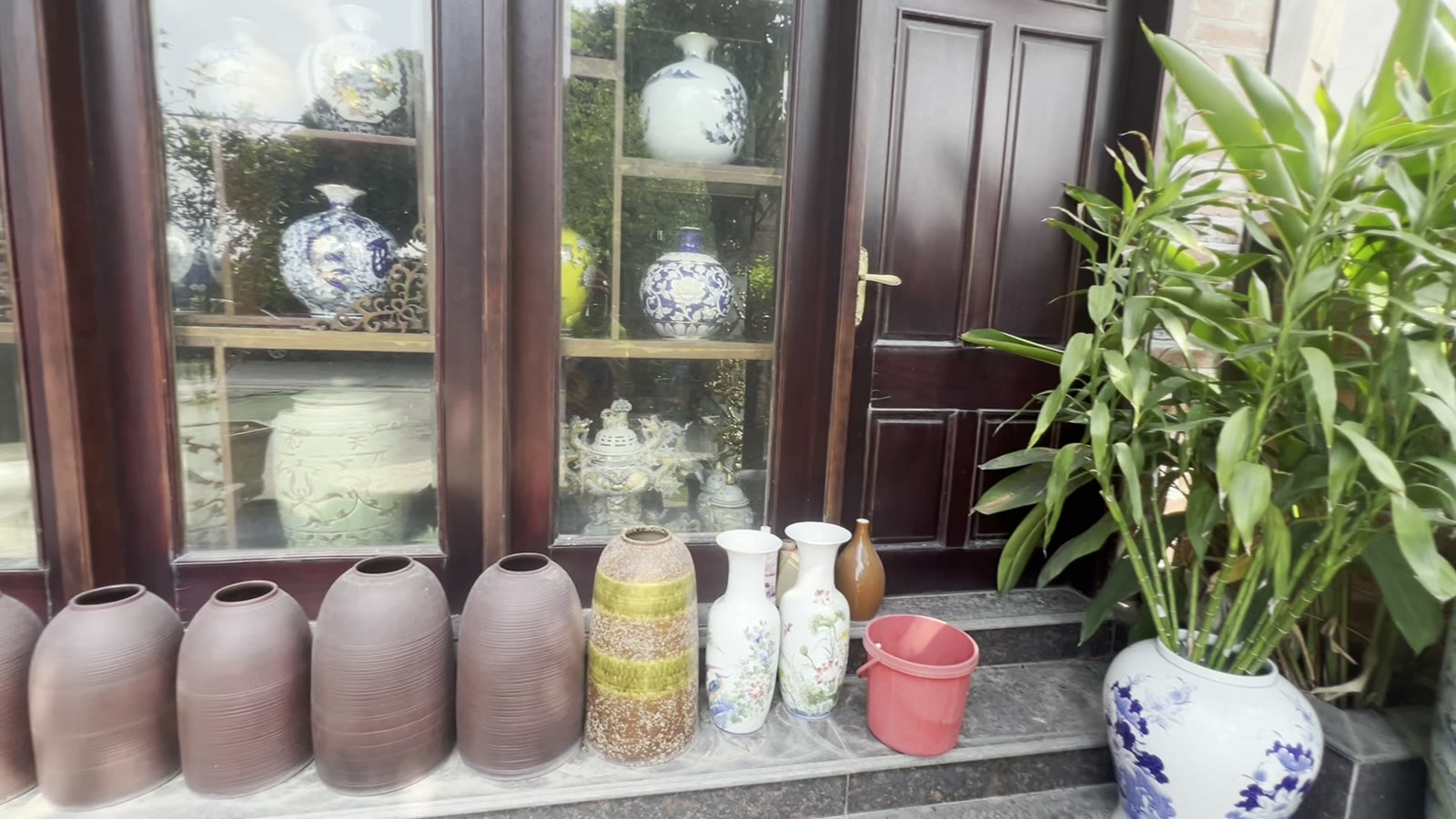 Tủ kính nhà gỗ trưng bình men lam, trước cửa bày bình gốm nâu và bình hoa sứ